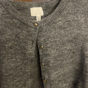 Grå stickad kofta med guldfärgade knappar från H&M - Mysig grå stickad kofta från H&M med rund halsringning och snygga guldfärgade knappar framtill. Perfekt att slänga på sig när det är lite kyligt ute. Klassisk modell som passar till det mesta.