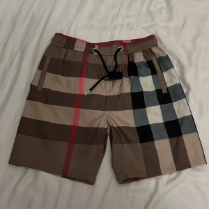  badshorts från Burberry - Säljer ett par klassiska badshorts från Burberry med det ikoniska rutiga mönstret i beige, svart, vitt och rött. Shortsen har snörning i midjan och en bakficka. Perfekta för sommaren och strandhäng!