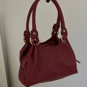 Vinröd handväska med gulddetaljer - Säljer en vinröd handväska med två rejäla handtag och snygga guldfärgade metalldetaljer. Väskan har dragkedja upptill och två fack med knapp på varsin sida. 