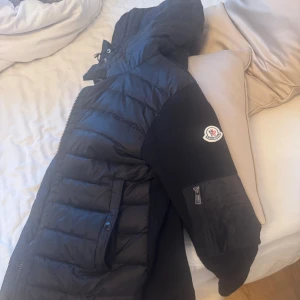 Svart jacka från Moncler - Säljer en svart pufferjacka från Moncler med huva och dragkedja. Moncler-logga på ärmen. Fickor och en extra ficka på ärmen. Perfekt för kyliga dagar. Storlek S