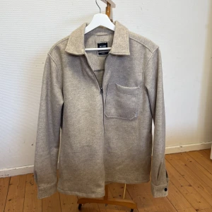 Beige overshirt från Only & Sons - Säljer en stilren beige overshirt från Only & Sons med dragkedja och bröstficka. Jackan har klassisk krage och långa ärmar, perfekt för lager-på-lager. Enkel och clean design som passar till mycket.
