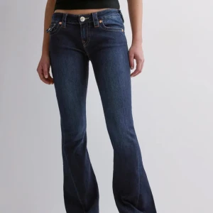 Mörkblå bootcut jeans från True Religion - Säljer ett par mörkblå jeans från True Religion med bootcut passform. Jeansen har kontrastsömmar, låg midja och detaljerade fickor med lock och knappar bak. Klassisk femficksmodell och silverfärgade knappar.