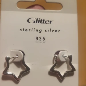 Stjärnformade örhängen i äkta silver från Glitter - Säljer ett par stjärnformade örhängen i sterling silver 925 från Glitter. Snygg och modern design som passar perfekt för dig som gillar unika smycken. Örhängena är i blankt silver och har en cool, öppen stjärnform.