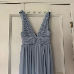 Ljusblå balklänning från Make Way - Säljer en elegant ljusblå långklänning från Make Way med v-ringad överdel och breda axelband. Klänningen har fina veckade detaljer över bysten och midjan samt dragkedja i ryggen. Perfekt för bal eller festliga tillfällen. Aldrig använd av mig men köpt på vinted. Har gått en tråd på baksidan (se sista bilden) men det är inget som syns. 