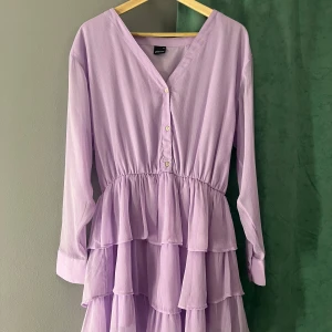 Ljuslila volangklänning med knappar - Säljer en ljuslila klänning med v-ringning, långa ärmar och volanger i flera lager. Jag har använt klänningen endast 1 gång och det syns inga tecken på användning🥰