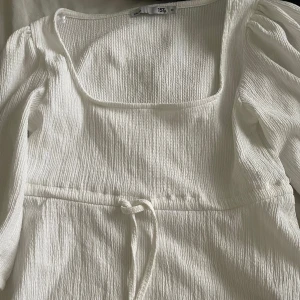 Vit blus med knyt i midjan - Säljer en söt vit blus med fyrkantig ringning och knytband i midjan. Blusen har puffiga trekvartsärmar och är gjord i ett lätt, strukturerat material som ger en luftig känsla. Perfekt för varma dagar!