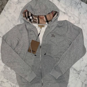 Klassisk grå burberry zip - Snygg grå hoodie från Burberry med dragkedja och huva. Insidan är fodrad med klassiskt Burberry-rutigt tyg. Broderad logga på bröstet och ribbade muddar. Perfekt för en stilren och bekväm look.