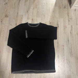 Svart stickad tröja med vita detaljer - Svart stickad tröja med vita dekorativa sömmar vid halsringning, ärmslut och nederkant. Klassisk rund hals och lång ärm. Perfekt för dig som gillar stilrena plagg med en twist.