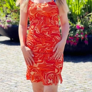 Orange mönstrad kortklänning - Säljer en färgstark orange kortklänning med abstrakt mönster i olika nyanser av orange och rött. Klänningen är ärmlös och har en figurnära passform med volangdetalj nertill. Perfekt för varma dagar!