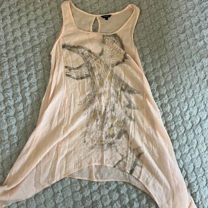 Beige linnetopp med drakemotiv Lindex - Säljer en supertunn och luftig beige linnetopp från Lindex i storlek M. Toppen har ett coolt motiv framtill i gråa och gula toner, är ärmlös och har en asymmetrisk, flowy nederkant. Perfekt för varma dagar när du vill sticka ut lite extra.