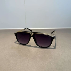Louis Vuitton svarta och guldiga solglasögon - Snygga solglasögon från Louis Vuitton med svart båge och guldiga detaljer. Modellen har en cool pilotform och mörka tonade glas. Sidorna pryds av Louis Vuitton-loggan och designen känns både lyxig och modern.