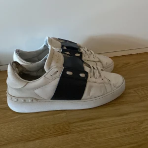 Valentino Garavani Open sneakers vit/blå - Snygga Valentino Garavani Open sneakers i vitt skinn med bred mörkblå rem över vristen och coola nitar på hälen. Klassisk rund tå, platt sula och snörning framtill. Perfekta för dig som vill ha en lyxig och stilren look med edge. Små defekter men de gör inte skorna sämre 