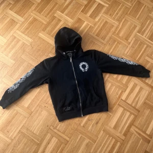 Svart Chrome Hearts hoodie med tryck - Svart hoodie från Chrome Hearts med stora vita tryck både fram och bak samt detaljerade mönster längs ärmarna. Dragkedja framtill, huva och fickor. Grym streetwear vibe och riktigt snygga detaljer. Perfekt för dig som gillar statement-plagg.
