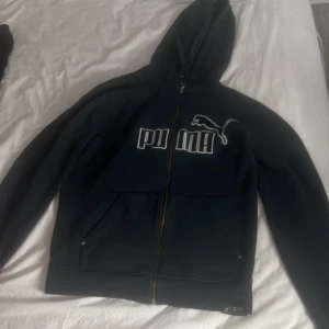 Svart hoodie från Puma med dragkedja - Svart hoodie från Puma med stor vit logga och text på bröstet. Tröjan har huva, hel dragkedja och två fickor framtill. Perfekt för en sportig och avslappnad stil.
