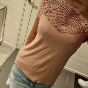 Beige blus med spets från H&M - Supersöt beige blus från H&M med detaljerad spets upptill och över axlarna. Toppen är ärmlös och har en mjuk, lätt figurnära passform. Perfekt för dig som gillar romantiska och feminina vibes. Säljes i storlek S.