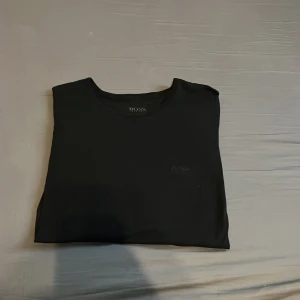 Svart t-shirt från Hugo Boss - En stilren svart t-shirt från Hugo Boss med diskret logga på bröstet. Klassisk rund halsringning och korta ärmar. Tillverkad i mjuk bomull som känns skön mot huden. Perfekt för dig som gillar en clean och enkel look. Hör av er vid minsta lilla fråga. 