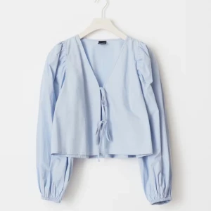 Ljusblå blus med puffärm och knyt - Supersöt ljusblå blus med v-ringning, puffiga långärmar och två knyt framtill. Croppad modell som ger en trendig siluett. Perfekt att styla med jeans eller kjol för en cool look. Helt ny med prislapp