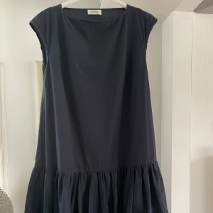 Mörkblå volangklänning från italienska Ottod'Ame - Snygg mörkblå klänning från Ottod'Ame i 100% bomull. Modellen är loose fit med volang nertill och ärmlös design. Perfekt för dig som gillar en enkel men trendig look med italiensk känsla.