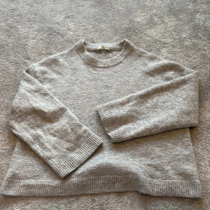 Grå stickad tröja från Gina Tricot - Mysig grå stickad tröja från Gina Tricot i premiumkvalitet. Tröjan har en klassisk rund halsringning, långa ärmar och ribbade muddar vid ärmslut och nederkant. Perfekt för lager-på-lager och chill dagar.