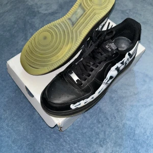 Nike Air Force 1 Skeleton svart/vit - Billigaste på plick! Unika Nike Air Force 1 Skeleton i svart skinn med vit skelettgrafik längs sidorna. Klassisk låg modell med snörning, rund tå och genomskinlig sula. Sulan lyser på natten när man går! Perfekta för dig som vill sticka ut med en fet och edgy sneaker. Köpta för 7000kr på farfetch. Äkta låda till de följer med.