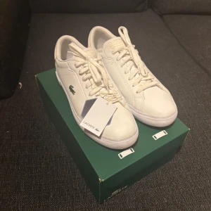 Vita sneakers från Lacoste - Snygga vita sneakers från Lacoste med klassisk låg profil och grönt krokodilmärke på sidan. Skorna har snörning, rund tå och är tillverkade i skinn med detaljer i textil vid hälen. Perfekta för en clean och stilren look. Helt nya, pris kan diskuteras vid snabb affär☺️