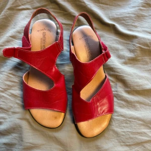 Röda sandaler i skinn från Remedios Miralles - Snygga röda sandaler i mjukt skinn med coola utskärningar och justerbar kardborreknäppning. De har öppen tå, platt sula och svart yttersula. Perfekta för varma dagar när du vill ha både stil och komfort.
