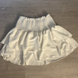 Vit volangkjol från SHEIN - Supersöt vit kortkjol från SHEIN med bred smock i midjan och dubbla volanger. Kjolen är gjord i 100% polyester och har en lätt, flowy känsla som passar perfekt till varma dagar. Elastisk midja för extra komfort och snygg passform. Aldrig använd. 