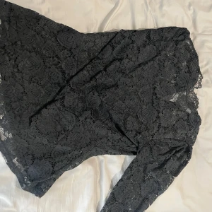 Svart blus med lång ärm - Supersnygg svart blus i spets med långärmade armar och figurnära passform. Blusen har ett vackert blommigt spetsmönster och är perfekt för dig som vill sticka ut med en elegant och trendig look.