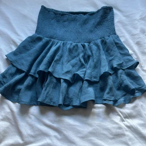 Blå volangkjol - Supersöt blå kortkjol med bred smock i midjan och dubbla volanger. Kjolen har en luftig känsla och är knappt använd. Här av dog vid prosförsöag pch frågor!