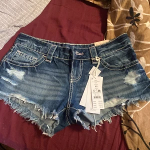 Lågmidjade jeansshorts från Gina Tricot - Snygga lågmidjade jeansshorts från Gina Tricot i mellanblå denim. Modellen har slitna detaljer, fransig kant och klassiska fickor både fram och bak. Perfekta för en chill sommarlook och passar till allt från sneakers till sandaler.