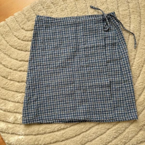 Blårutig omlottkjol från Hennes - Supersöt kort omlottkjol från Hennes i blått och vitt rutigt mönster. Kjolen har knytband i midjan och är gjord i mjuk bomull, perfekt för en chill och avslappnad look. Passar dig som gillar retrovibbar och vill ha något lättburet till sommaren.