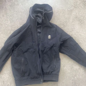 Svart Nike Air Max vindjacka med huva - Svart Nike Air Max vindjacka med huva och dragkedja framtill. Jackan har två fickor med dragkedja, reflekterande detaljer vid huvan och stor AIR MAX-logga på ryggen. Perfekt för dig som gillar sportig och clean stil.