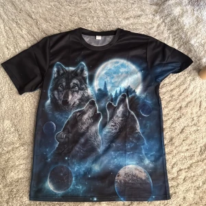 Svart t-shirt med vargar och måne - Cool svart t-shirt med ett stort tryck av tre vargar som ylar mot en blå fullmåne och rymdmotiv. T-shirten har korta ärmar och är gjord i ett glansigt, mjukt material som känns sportigt. Perfekt för dig som gillar unika och mystiska motiv. Denna e så kallad ”Alpha tröja”
