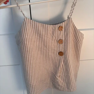 Beige och vit randig linnetopp - Säljer en beige och vit randig linnetopp med smala axelband och tre snygga träknappar framtill. Toppen har en rak passform och är perfekt för varma dagar. 