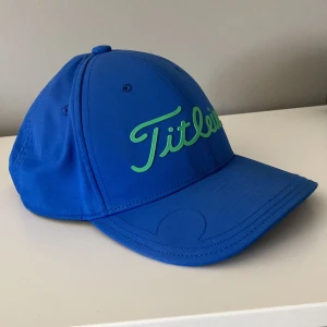 Blå Titleist keps  - Snygg blå keps från Titleist med justerbar rem bak och grön logga framtill. Klassisk böjd skärm och ventilerande hål upptill. Perfekt för golfbanan. Tillverkad i lätt och bekvämt material. Ny pris 500 mitt pris 189