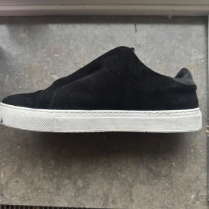 Axel Arigato - Snygga svarta sneakers från Axel Arigato i mocka med vita platta sulor. Skorna har dragkedja istället för klassisk snörning, vilket ger en unik och modern look. Perfekta för dig som gillar stilrena och minimalistiska skor med en twist.