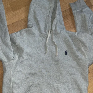 Grå hoodie från Polo Ralph Lauren - Snygg ljusgrå hoodie från Polo Ralph Lauren med klassisk blå logga broderad på bröstet. Tröjan har huva med snörning, magficka och långa ärmar. Perfekt för en chill och stilren look.