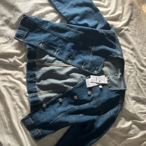Blå jeansjacka från mind: i storlek M - Snygg blå jeansjacka : i klassisk modell med rund halsringning och silvriga knappar framtill. Jackan har långa ärmar och markerade sömmar som ger en clean look. Perfekt att slänga över en hoodie eller t-shirt för en chill vibe.