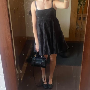 Svart kort klänning med smock - Supersöt svart kort klänning med smockad överdel och tunna axelband. Kjolen är luftig och har en lätt volangig känsla. Perfekt för varma dagar och enkel att styla med accessoarer. Materialet känns mjukt och bekvämt mot huden.
