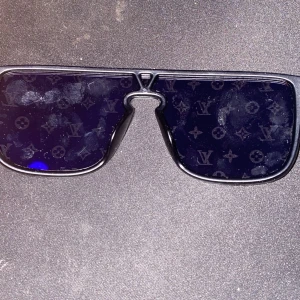 Svarta Louis Vuitton solglasögon - Snygga svarta solglasögon från Louis Vuitton med bred, rektangulär form och mörka linser. Linserna har LV-monogram över hela ytan för en lyxig vibe. Bågarna är svarta och robusta, perfekt för dig som vill sticka ut med en exklusiv accessoar.