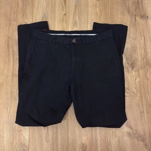 Svarta chinos från Cutter & Buck - Svarta chinos från Cutter & Buck med klassisk passform. Byxorna har snedställda fickor fram, bakfickor med knapp och bälteshällor. Tillverkade i bomull för en skön känsla och stilren look. Perfekta för dig som gillar en clean och enkel stil.