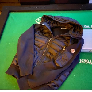 Moncler pufferjacka med huva - Säljer en  pufferjacka från Moncler med huva och dragkedja. Jackan har quiltade partier, ribbade muddar och en ficka med dragkedja på ärmen. Perfekt för dig som vill ha en stilren och varm jacka med snygga detaljer.