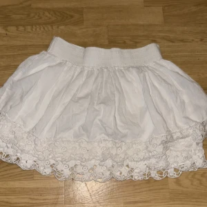 Vit kortkjol med spets från Gina Tricot - Superfin vit kortkjol från Gina Tricot med bred resår i midjan och volangig form. Kjolen har vacker spetsdetalj längst ner med blommigt broderi som ger en romantisk vibe. Perfekt för sommarens alla tillfällen!
