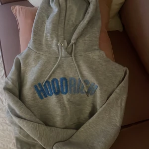 Grå Hoodie från Hoodrich med blå text - Säljer en grå hoodie från Hoodrich med broderad blå text framtill. Tröjan har huva med snörning och långa ärmar. 