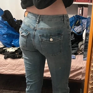 Blå bootcut jeans med ficklock - Snygga blå jeans från Gina Tricot med bootcut-ben och klassisk femficksdesign. Bakfickorna har lock och silverfärgade knappar. Jeansen är i denim med en något ljusare tvätt och har bälteshällor i midjan.