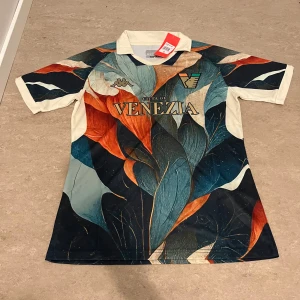 Venezia FC x Kappa matchtröja - Unik Venezia FC fotbollströja från Kappa med färgstarkt bladmönster i blått, orange, beige och svart. Tröjan har klassisk krage, korta ärmar och tryckt klubbmärke samt Kappa-logga på bröstet. Tillverkad i lätt och ventilerande material.