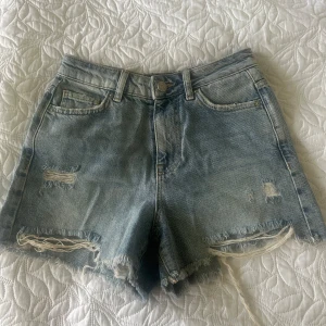 Blå jeansshorts med slitningar från Pieces - Snygga blå jeansshorts från Pieces i klassisk femficksmodell. Shortsen har råa kanter och slitna detaljer framtill och baktill för en avslappnad vibe. Perfekta för sommaren och lätta att matcha med allt. Stängs med knapp och dragkedja.
