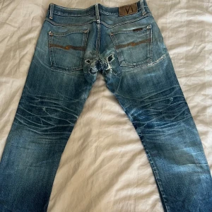 Nudie Jeans blå selvedge - Säljer ett par blå jeans från Nudie Jeans med tydliga slitningar och lagningar på både fram- och baksida. Klassisk femficksmodell med orange sömmar och Nudie-logga på bakfickan. Jeansen har en straight fit och är tillverkade i denim.