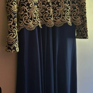 Blå långklänning med guldmönster Famelin - Elegant långklänning från Famelin i djupblått tyg med lyxigt broderat guldmönster upptill och på ärmarna. Klänningen har lång ärm och vackert fall. Perfekt för dig som vill sticka ut med en klassisk och glamorös look.