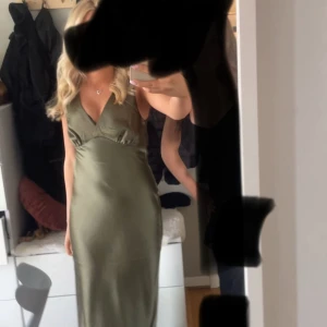 Olivgrön långklänning i satin - Säljer en olivgrön långklänning i satin med djup v-ringning och ärmlös design. Klänningen har en figurnära passform och ett elegant, slätt fall som ger en lyxig känsla. Perfekt för dig som vill sticka ut med en stilren och trendig look.